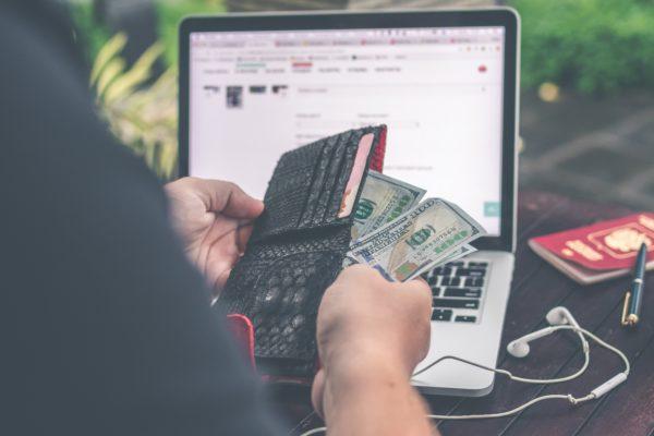 Man holding wallet and cash, sitting at his laptop - travel prep costs